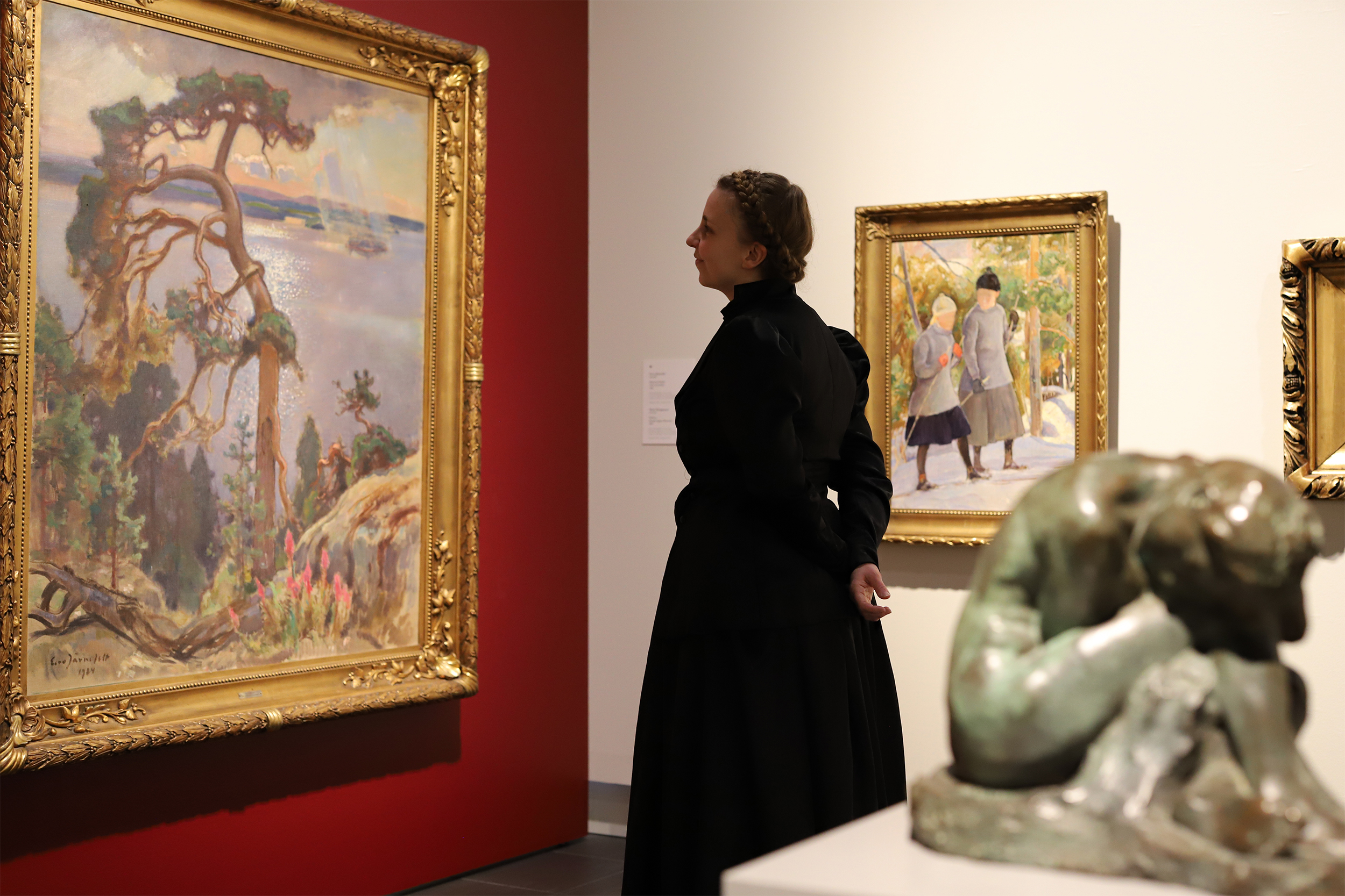 Taidemuseon näyttelytila, jonka keskellä seisoo vanhaan juhlavaan mekkoon pukeutunut nainen katsellen suurikokoista maisemamaalausta.